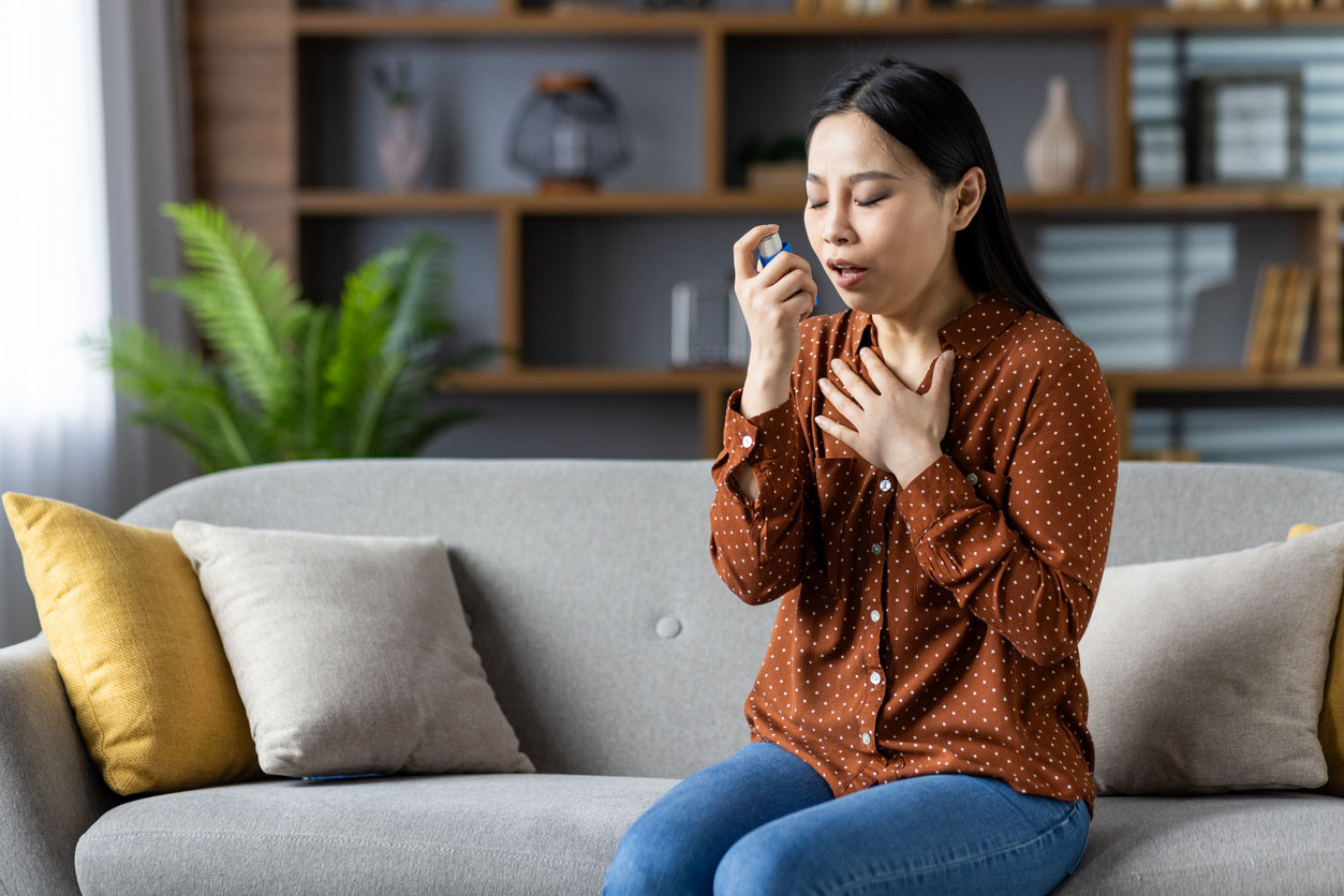 Asian woman using asthma inhaler, sitting on sofa experiencing breathing issue. Wearing polka dot blouse, managing respiratory health at home. Focused, calming atmosphere.