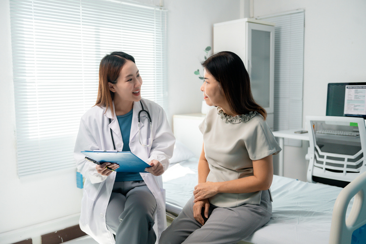 Female physician reviewing medical chart while conversing with seated senior patient inside contemporary healthcare facility, discussing diagnostic details and potential treatment strategies