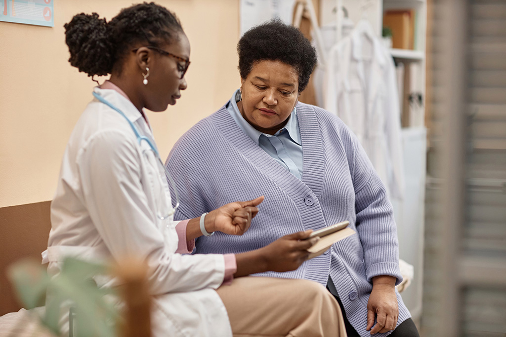 Senior African American woman looking at tablet in hand of female doctor talking about test results in clinic office