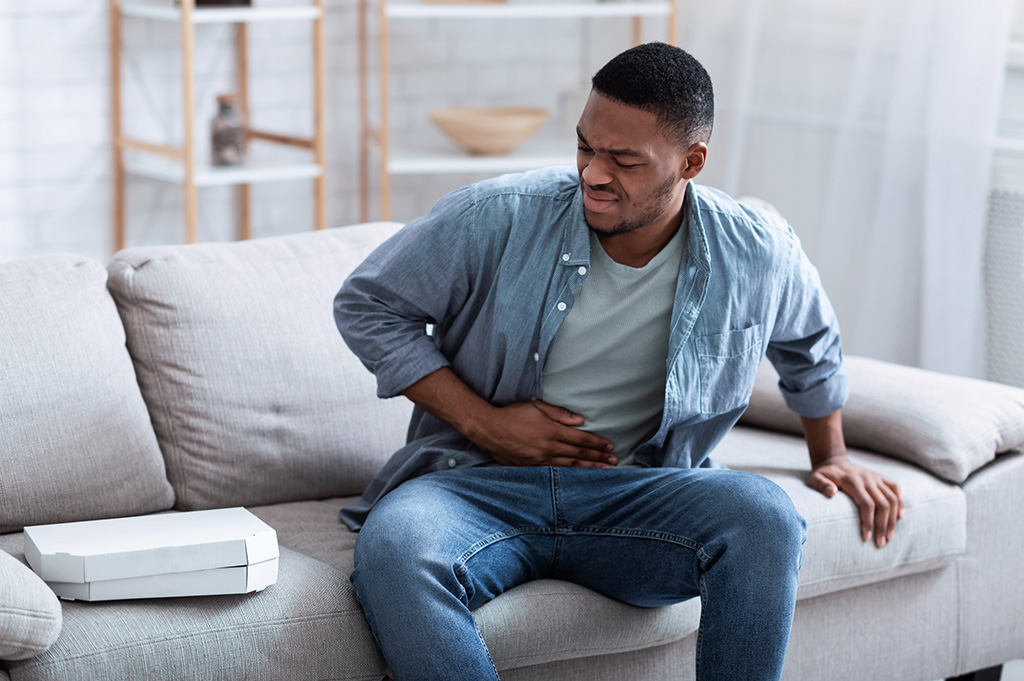 African American Guy Having Stomachache After Eating Pizza Touching Aching Stomach Suffering From Pain Sitting On Sofa At Home. Food Poisoning, Gastritis, Abdominal Inflammation And Appendicitis.