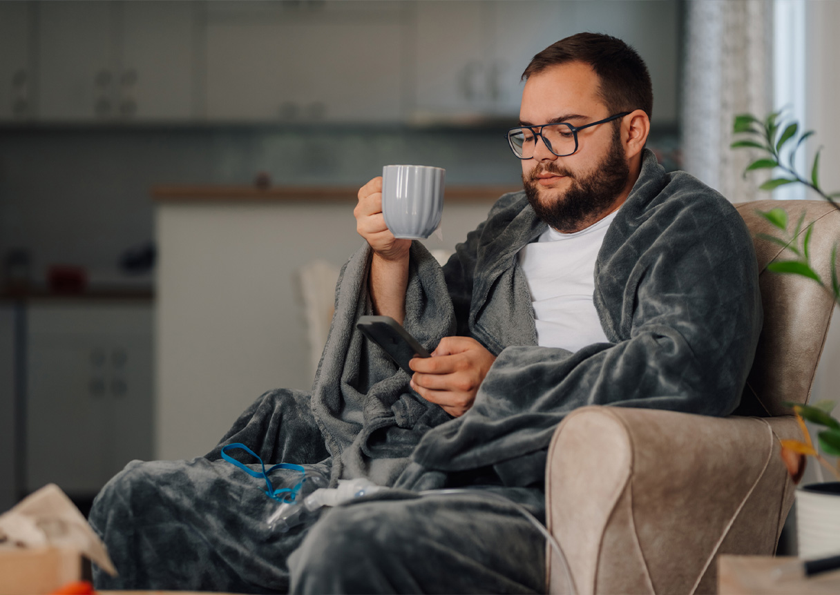 Bearded man with the flu, wearing bathrobe, sitting on sofa, drinking tea while on a telemedicine call, with nebulizer mask near him