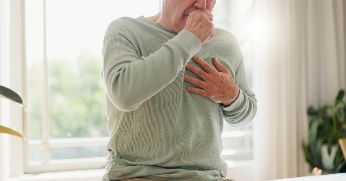 A person sitting on a sofa holding their chest in pain while coughing, illustrating the warning signs of a heart attack or cardiac distress.
