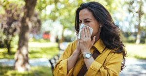 A woman in a yellow top blowing her nose with a tissue, depicting common cold or allergy symptoms treated at AllCare.