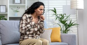 A sick woman in a sweater coughing while sitting on her couch at home, highlighting when to seek care for persistent coughs at AllCare.