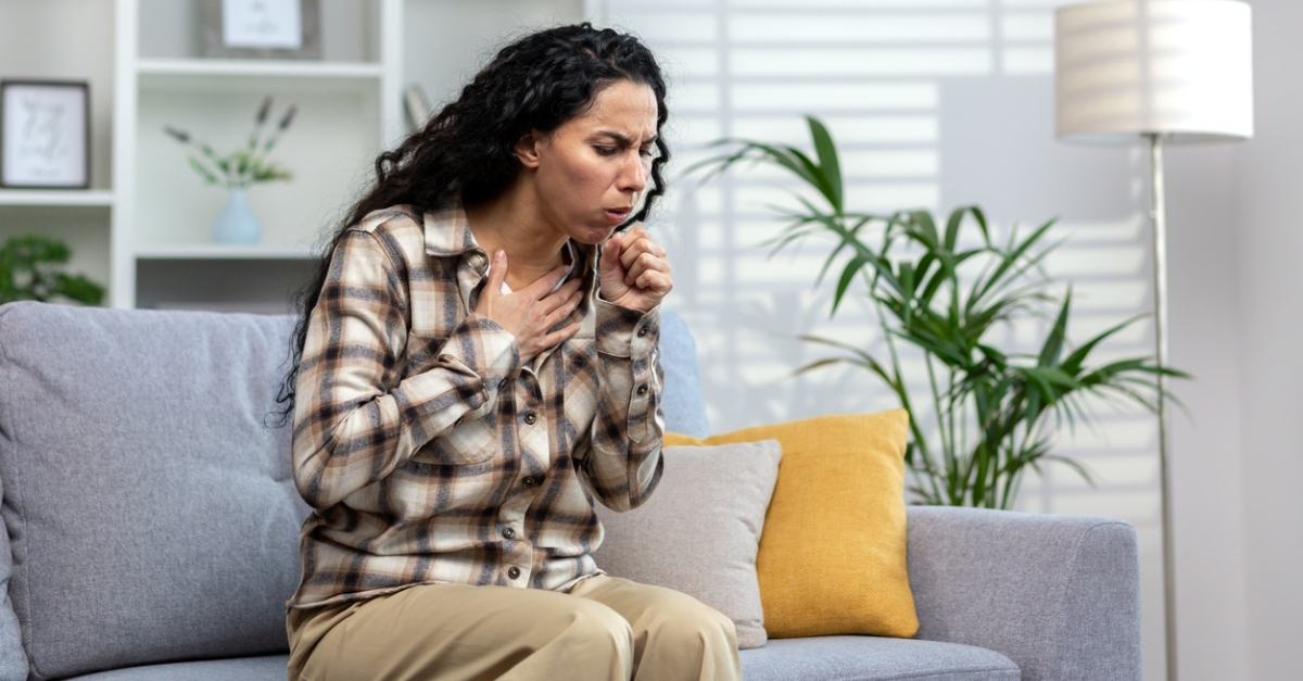 A sick woman in a sweater coughing while sitting on her couch at home, highlighting when to seek care for persistent coughs at AllCare.